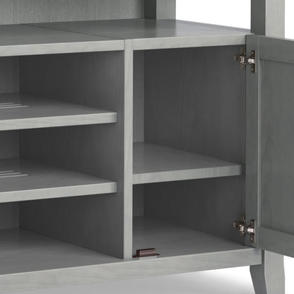 The Amherst TV Media Stand, handcrafted in gray wood, features an open cabinet with two interior shelves, metal door hinges, and a small brown rectangular object on the bottom shelf.