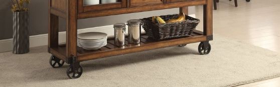The Kadri Kitchen Cart in Distressed Chestnut with black metal wheels holds plates, glass jars, and a black fruit basket on a beige rug in a room with wooden floors and gray walls.