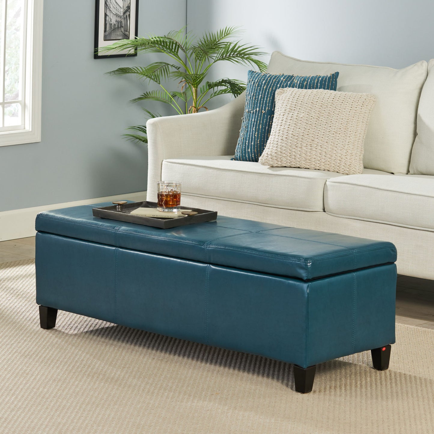 A modern living room featuring a beige sofa, textured pillows, and the Glouster - Contemporary Storage Ottoman in blue used as a coffee table with a tray and drink on top, plus a plant and framed picture against a pale blue wall.