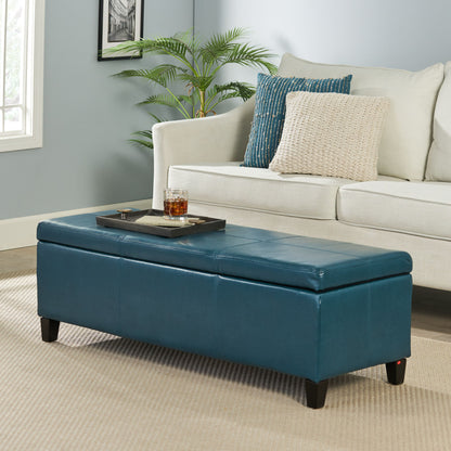 A modern living room featuring a beige sofa, textured pillows, and the Glouster - Contemporary Storage Ottoman in blue used as a coffee table with a tray and drink on top, plus a plant and framed picture against a pale blue wall.