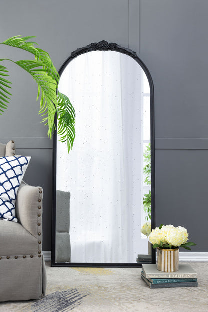 The Hand Carved Rose Antique Mirror Frame, a wood arch mirror wall decor, leans against a gray wall near a green plant, beige armchair with blue-and-white pillows, stacked books, and a vase of white flowers.