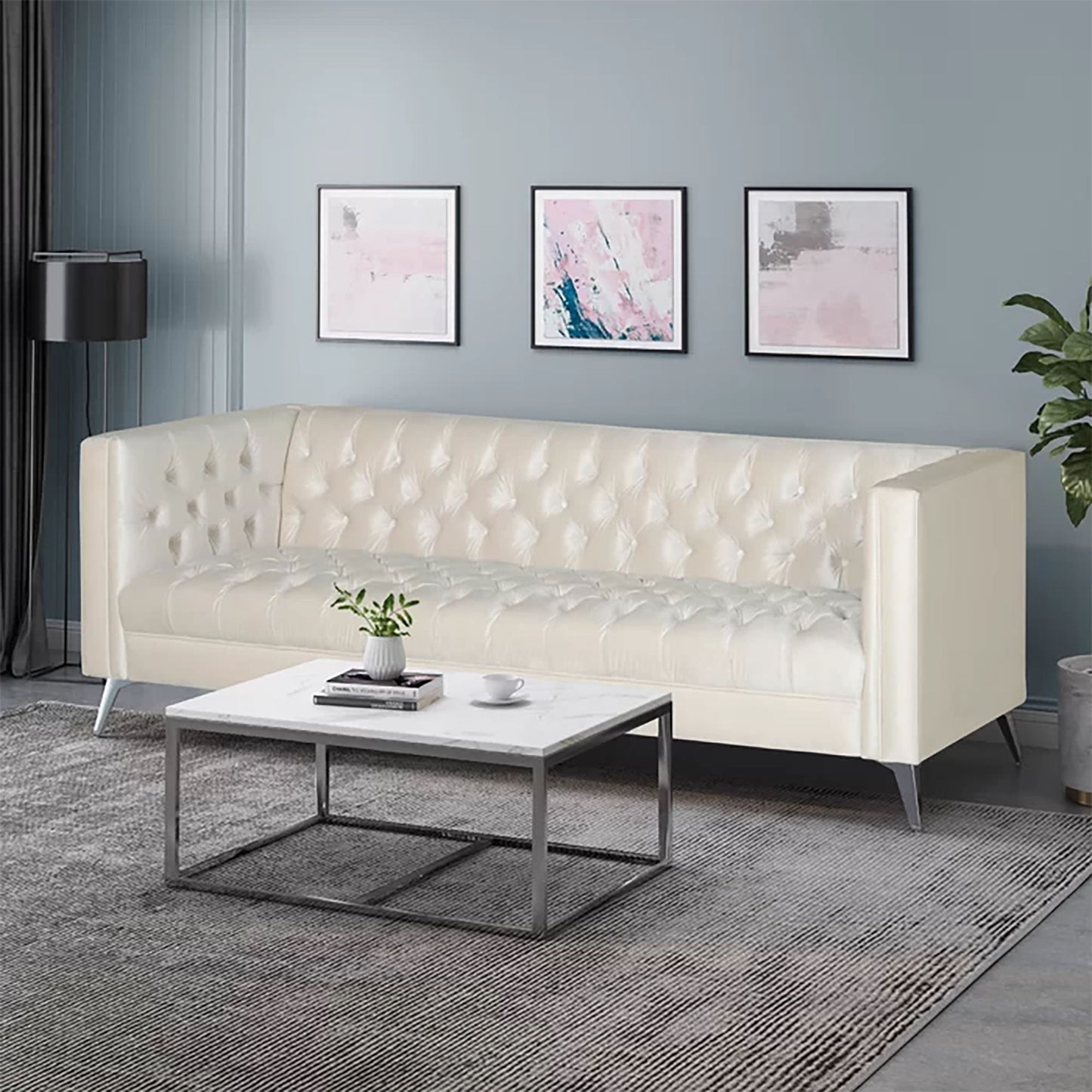 A modern living room featuring the Contemporary 3 Seater Sofa With Button Tufting in cream, a white marble coffee table with a plant and books, gray rug, three abstract pink and gray wall art pieces, and a black floor lamp against blue-gray walls.
