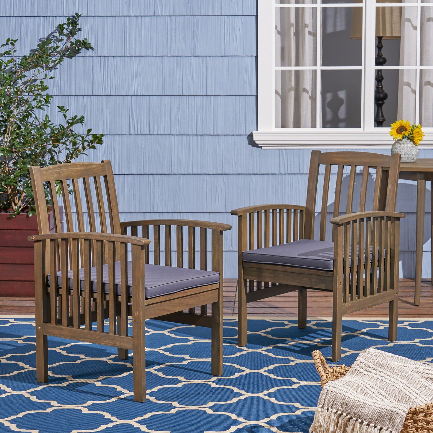 Two Casa - Acacia Dining Chair Set With Cushions, featuring gray cushions, sit on a blue patterned outdoor rug by a blue house with white trim. Nearby are a small table with a sunflower vase and a large potted plant.