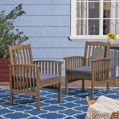 Two Casa - Acacia Dining Chair Set With Cushions, featuring gray cushions, sit on a blue patterned outdoor rug by a blue house with white trim. Nearby are a small table with a sunflower vase and a large potted plant.
