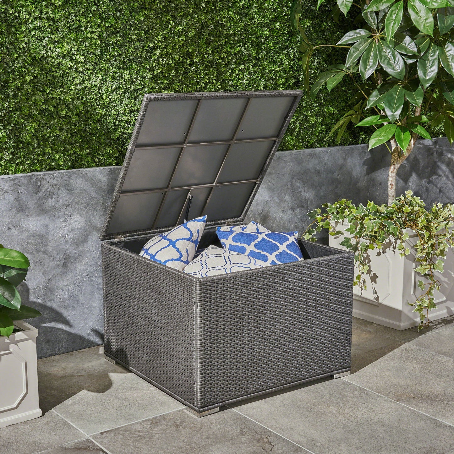 The Santa Rosa Storage Table, featuring a gray finish and hinged lid, is open on a stone patio with blue and white patterned cushions inside, surrounded by potted plants and a green hedge wall.