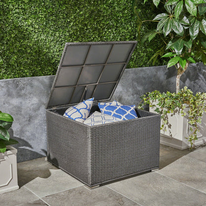 The Santa Rosa Storage Table, featuring a gray finish and hinged lid, is open on a stone patio with blue and white patterned cushions inside, surrounded by potted plants and a green hedge wall.
