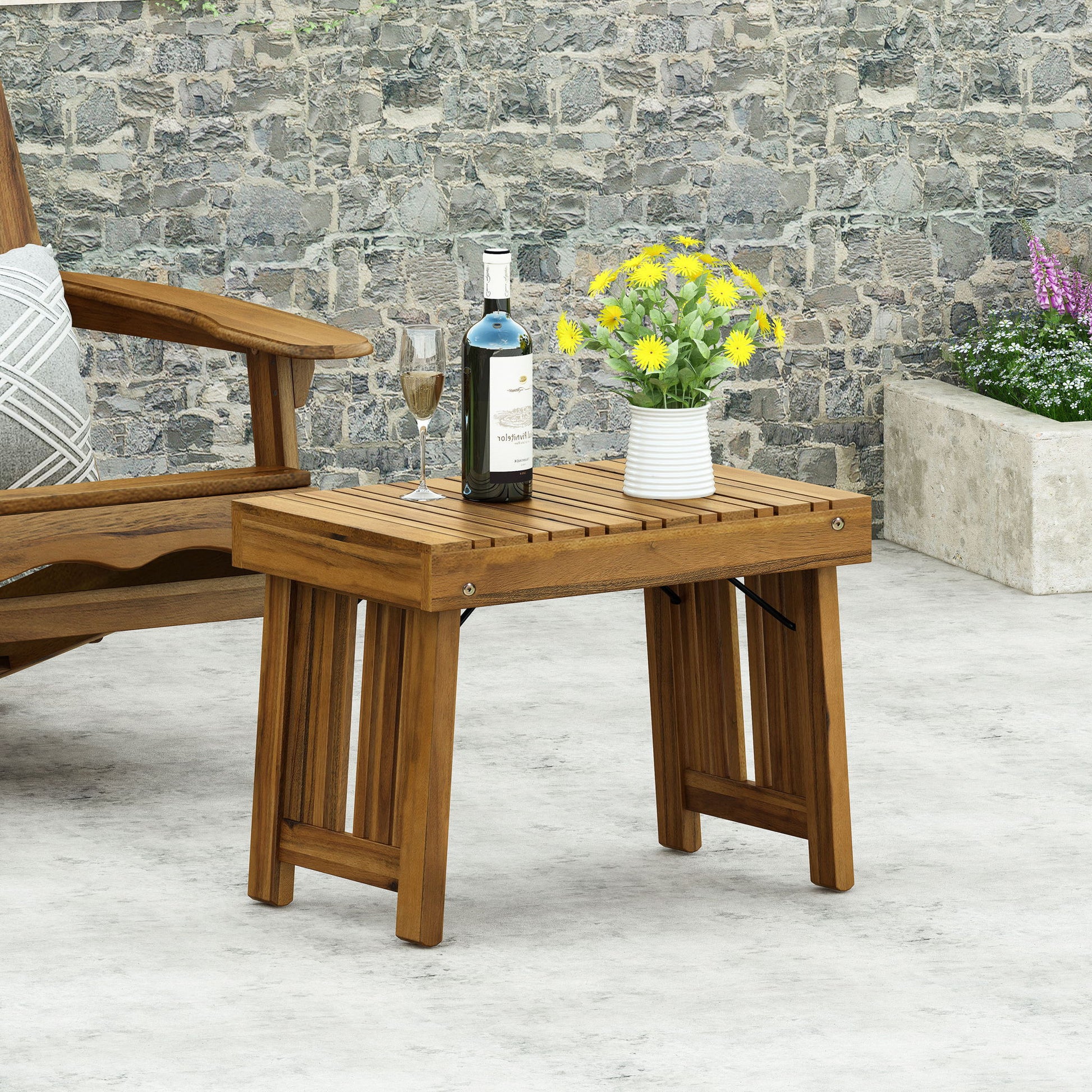 The Temptation Outdoor Foldable Side Table With Slat Design holds a bottle of wine, wine glass, and a white vase with yellow flowers. It's placed on a patio near a wooden chair and stone wall with greenery in the background.