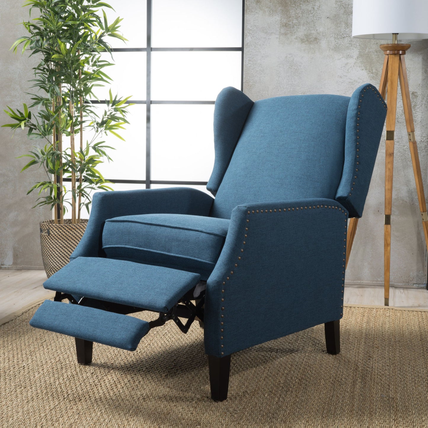 The Westeros Wing Back Fabric Upholstered Recliner, shown in blue with nailhead trim and extended footrest, sits on a woven rug with a potted plant, floor lamp, and textured walls in the background.