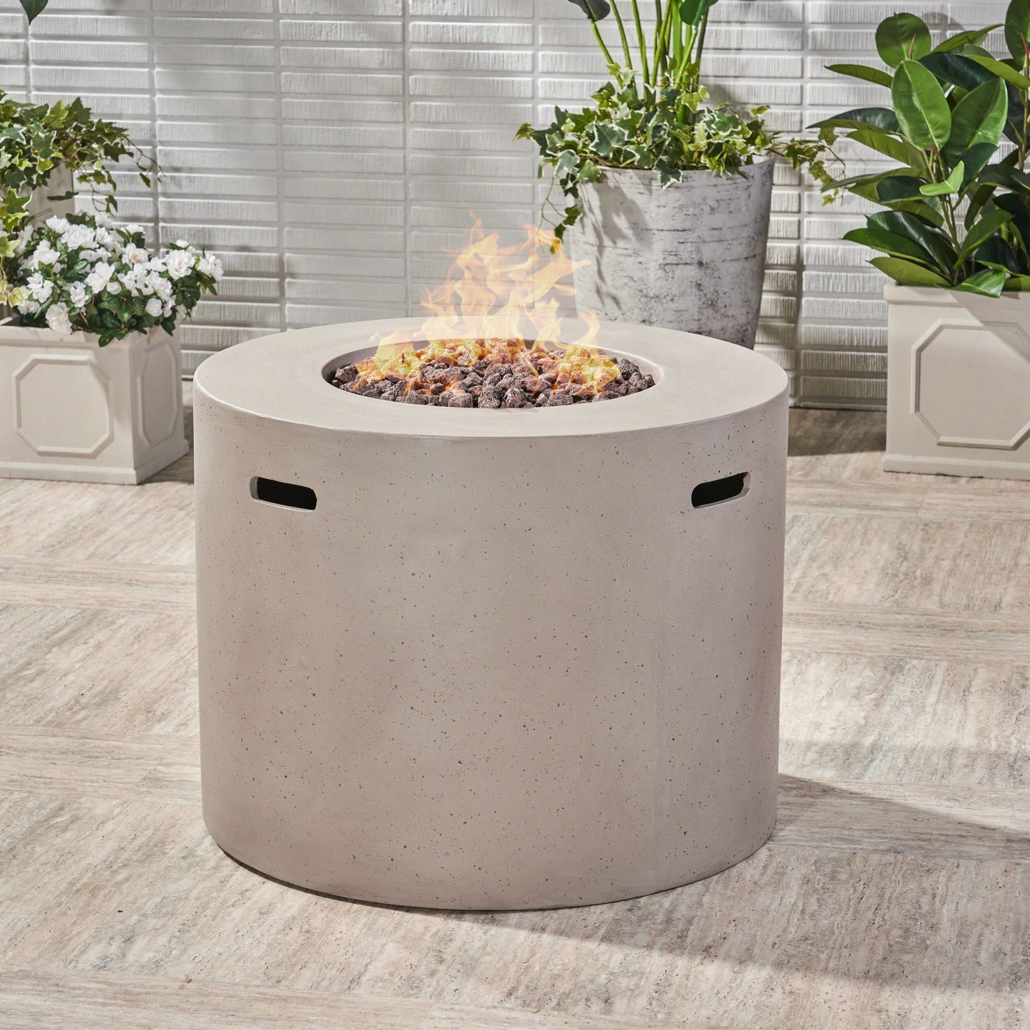 A modern Round Iron Fire Pit With 40,000 Btu burns at the center, surrounded by potted green plants against a tiled outdoor wall.
