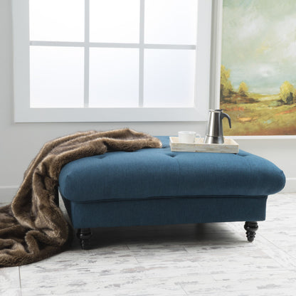 The Madrid - Tufted Ottoman Footrest, upholstered in plush blue fabric and draped with a fur throw, features a tray with a metal coffee pot and mug. It sits on a light wooden floor by a large window and a landscape painting.