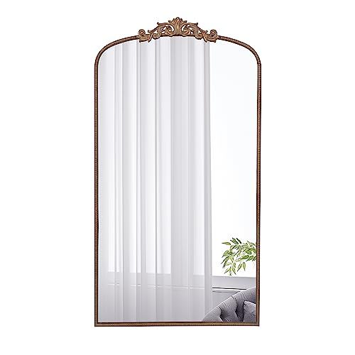The Arch Mirror, Baroque Inspired Wall Decor, features a tall rectangular shape with an ornate bronze frame and decorative crest, reflecting sheer white curtains, a window, green plant, and gray sofa. Ideal for bathroom, bedroom, or living room.
