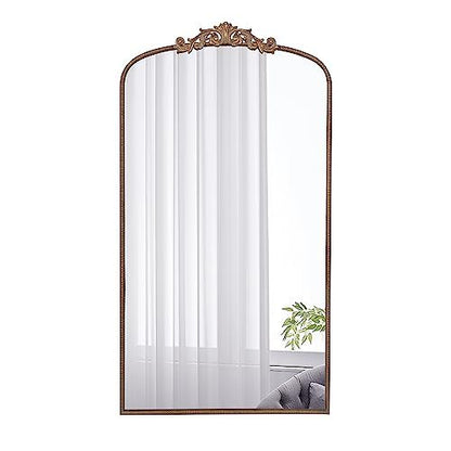 The Arch Mirror, Baroque Inspired Wall Decor, features a tall rectangular shape with an ornate bronze frame and decorative crest, reflecting sheer white curtains, a window, green plant, and gray sofa. Ideal for bathroom, bedroom, or living room.