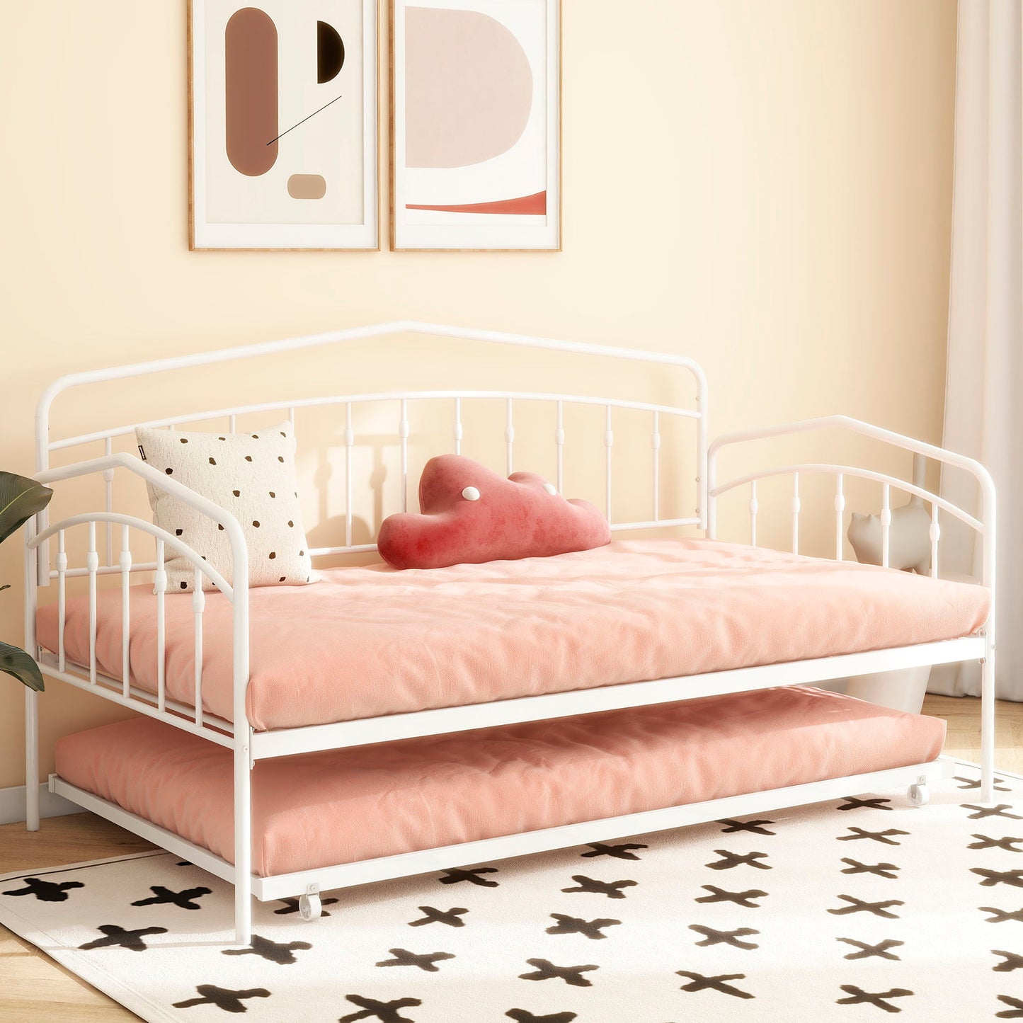 The Fox Daybed With Twin Trundle features white metal frame, pink bedding, a white pillow with black dots and a pink cloud-shaped pillow. It sits in a bright room with abstract wall art and a white rug adorned with black X patterns.