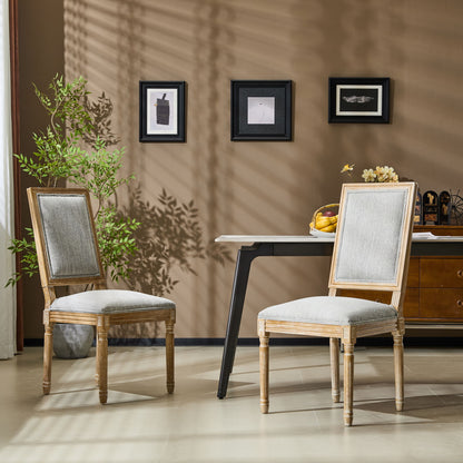 Two French Country Style Dining Chairs with upholstered seats and fluted legs are set in a modern room with a brown wall, three small framed pictures above, a desk with fruit and decor against the wall, and a green plant nearby.