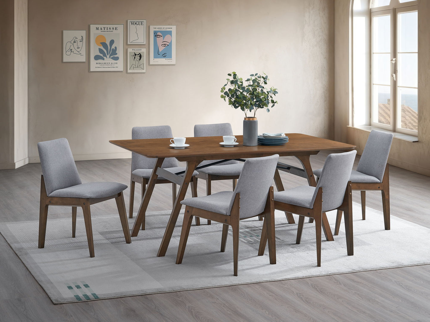 A modern dining room featuring the Kaela Dining Table in walnut, set for six with light gray upholstered chairs, a plant centerpiece, cups, and plates on a light rug. Minimalist wall art and large windows fill the space with natural light.