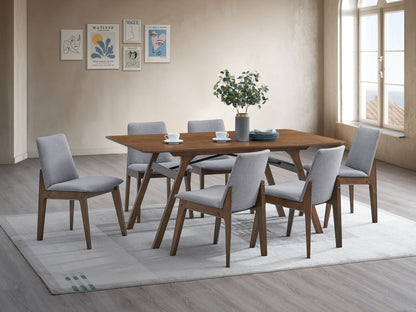 A modern dining room featuring the Kaela Dining Table in walnut, set for six with light gray upholstered chairs, a plant centerpiece, cups, and plates on a light rug. Minimalist wall art and large windows fill the space with natural light.