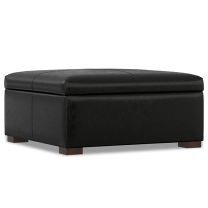 The Gabbie - Coffee Table Upholstered Storage Ottoman is a large, square black leather piece with subtle stitching and short wooden block feet, shown on a white background.