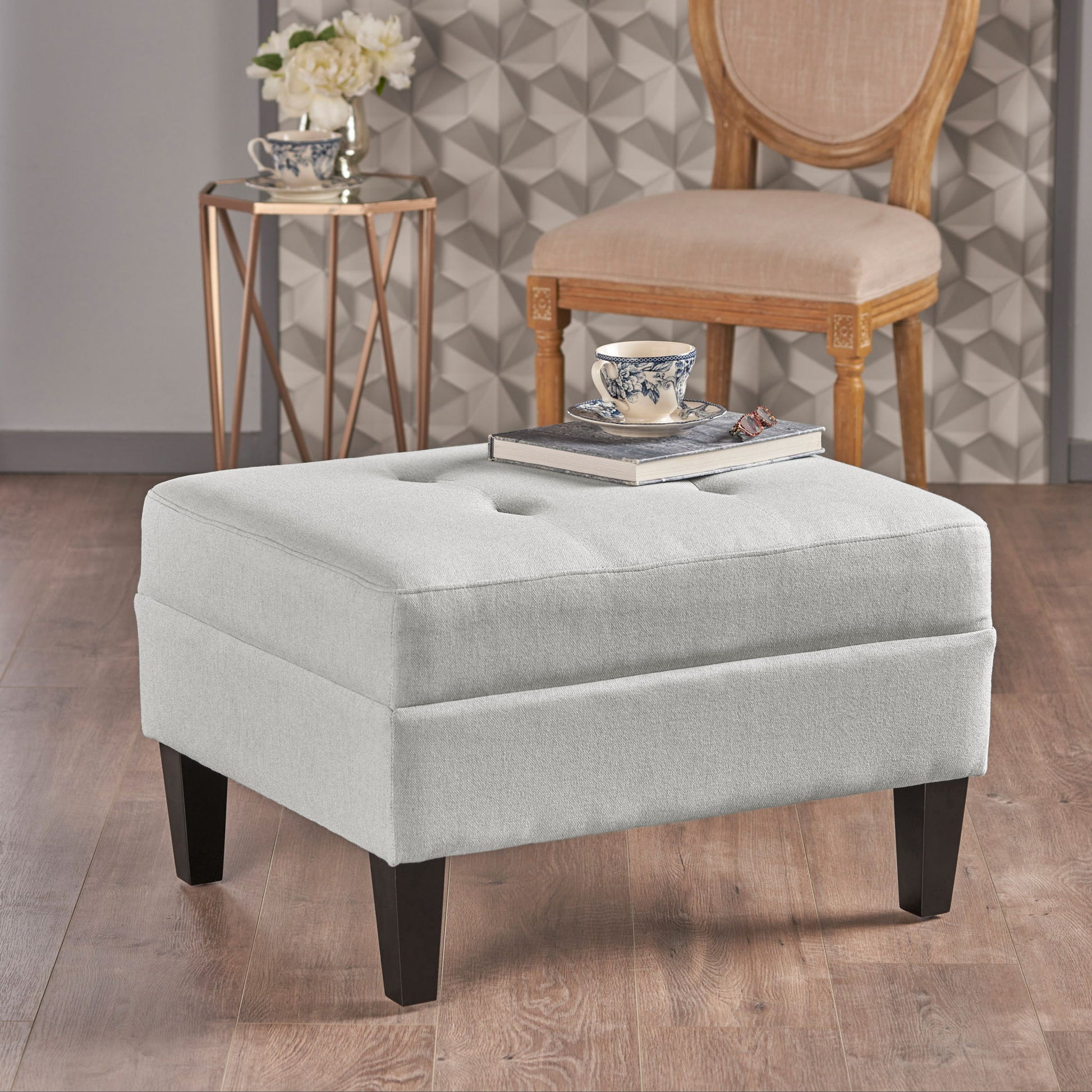 A Classic Ottoman With Birch Wood Legs And Fabric Upholstery, topped with a book and teacup, sits on a wooden floor. Behind it are a wooden chair and a small round table holding a flower vase and another teacup.