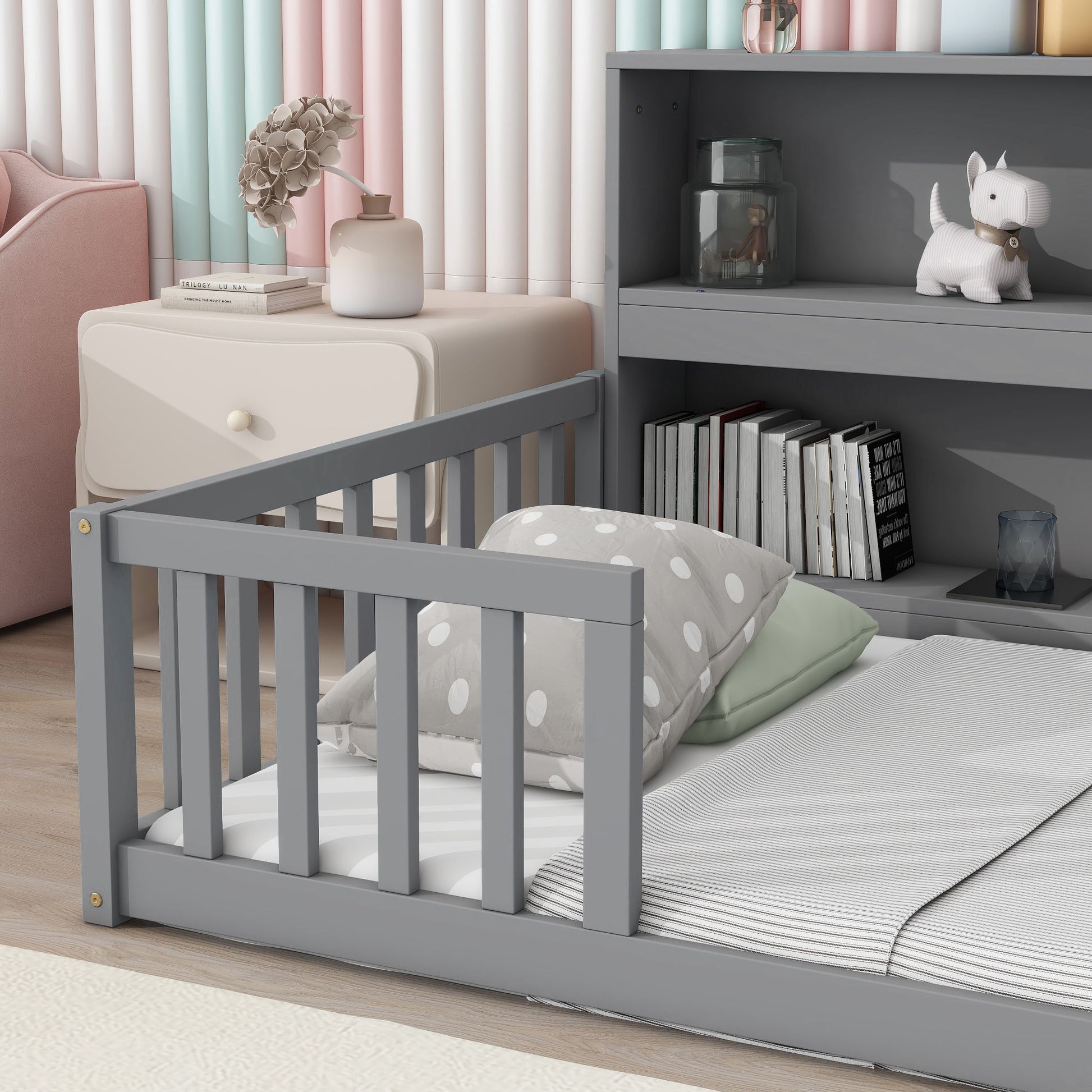 A child's bedroom featuring a gray Floor Bed with Bedside Bookcase, green and polka dot pillows, a nightstand with flowers and books, shelves with books, jars, a white dog figurine, and pastel-striped curtains in the background.