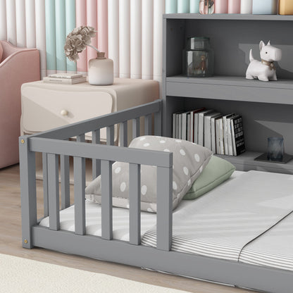 A child's bedroom featuring a gray Floor Bed with Bedside Bookcase, green and polka dot pillows, a nightstand with flowers and books, shelves with books, jars, a white dog figurine, and pastel-striped curtains in the background.