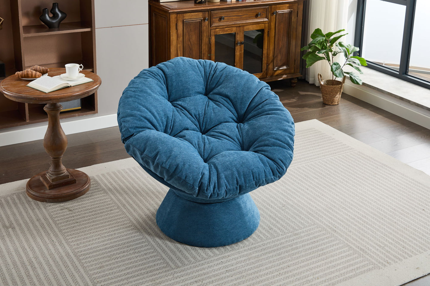 An Oversized Swivel Accent Chair in plush blue sits on a light patterned rug, surrounded by wood furniture, a round side table with books and a cup, and a potted plant near large windows in the living room.