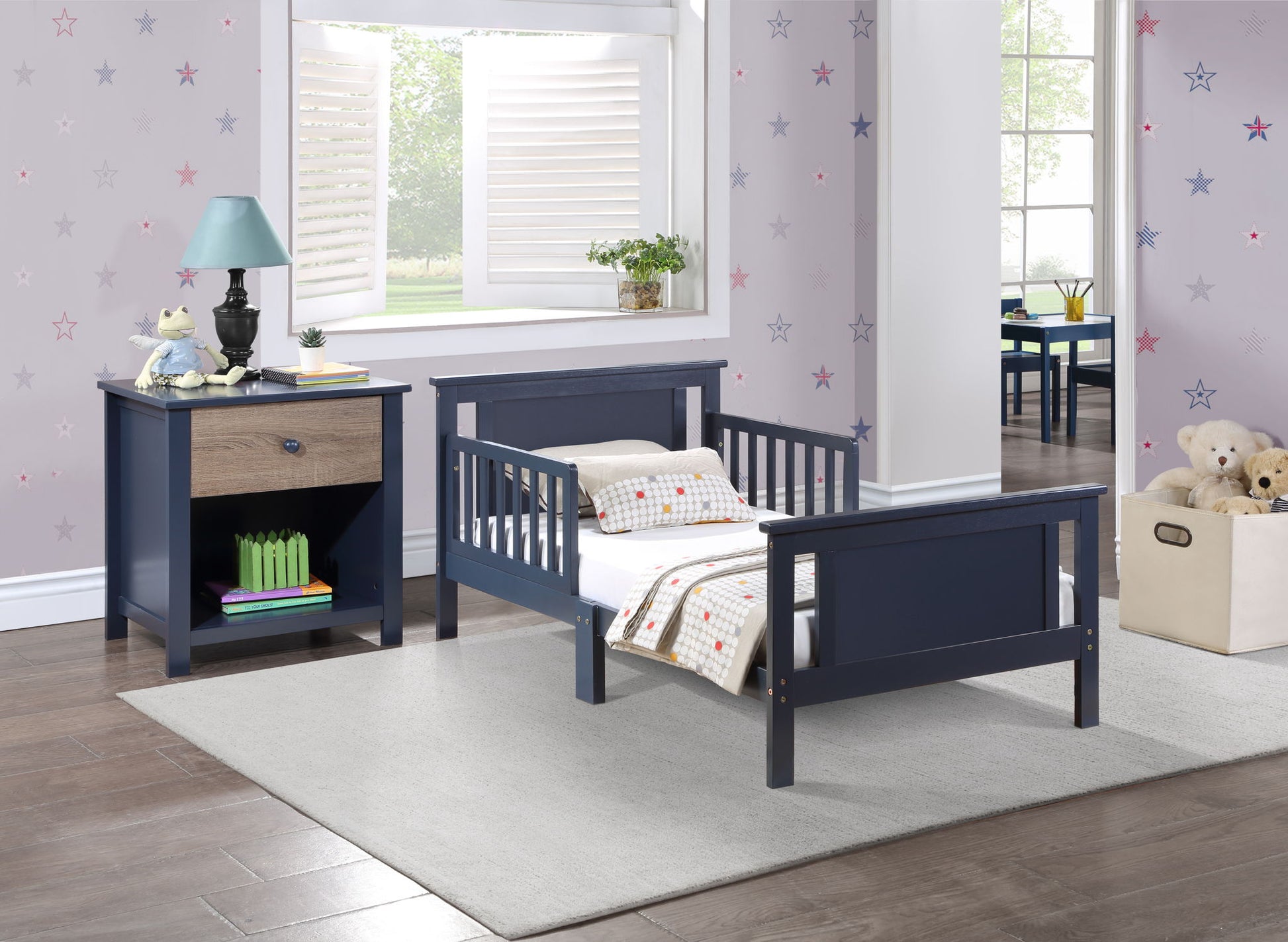 A cozy toddler bedroom features the Connelly Reversible Panel Toddler Bed in navy blue with a matching nightstand, a lamp and books, a soft rug, star-patterned walls, a toy bin in the corner, and a window overlooking a green yard.