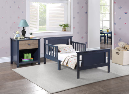 A cozy toddler bedroom features the Connelly Reversible Panel Toddler Bed in navy blue with a matching nightstand, a lamp and books, a soft rug, star-patterned walls, a toy bin in the corner, and a window overlooking a green yard.