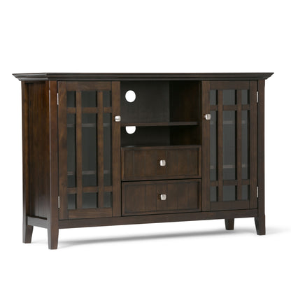 The Bedford Handcrafted Tall TV Media Stand features dark brown wood, glass-paneled doors on each side, two center open shelves with cable holes, two drawers below, four legs, and square metal handles.