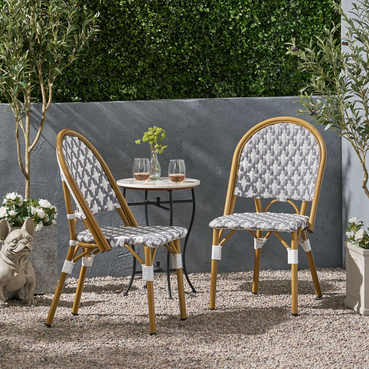 Two Chic French Bistro Chairs with durable rattan and aluminum frames sit by a small round table with glasses and a vase on a gravel patio, surrounded by potted plants, lush greenery, and a decorative dog statue.