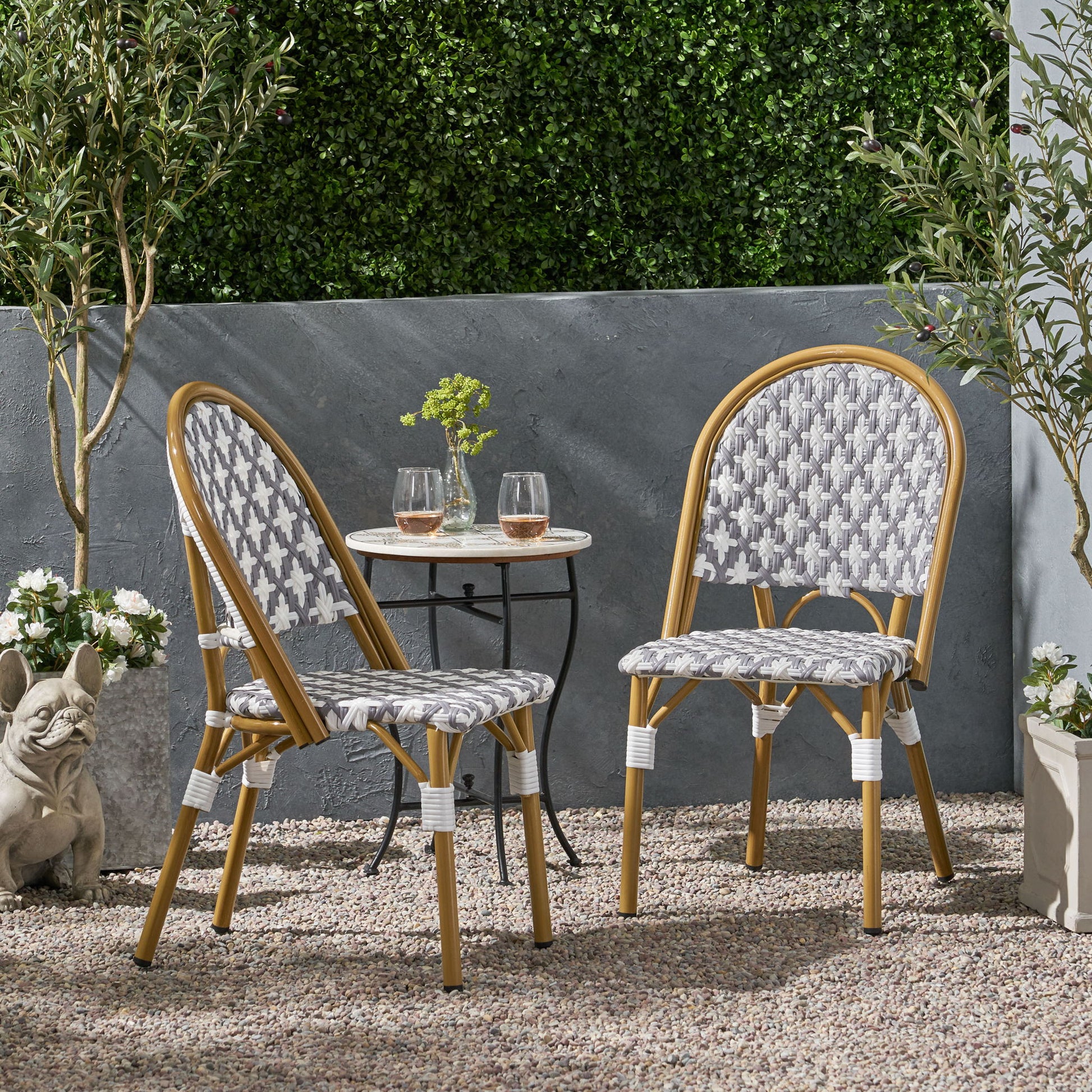 Two Chic French Bistro Chairs with durable rattan and aluminum frames sit by a small round table with glasses and a vase on a gravel patio, surrounded by potted plants, lush greenery, and a decorative dog statue.