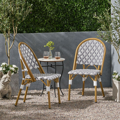 Two Chic French Bistro Chairs with durable rattan and aluminum frames sit by a small round table with glasses and a vase on a gravel patio, surrounded by potted plants, lush greenery, and a decorative dog statue.