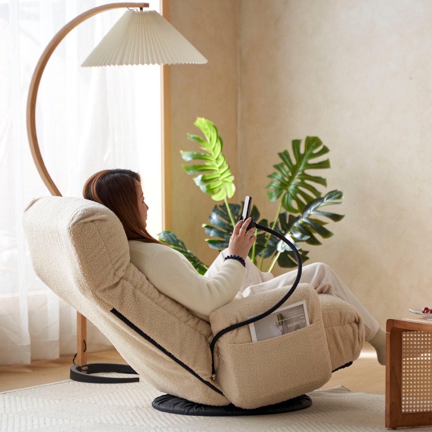 A woman relaxes in an Adjustable Swivel Lounge Chair With 360 Degree Rotating Seat And Headrest Support, checking her phone. Nearby are a side pocket with magazines, indoor plants, a floor lamp, and soft natural light in a calm living room.