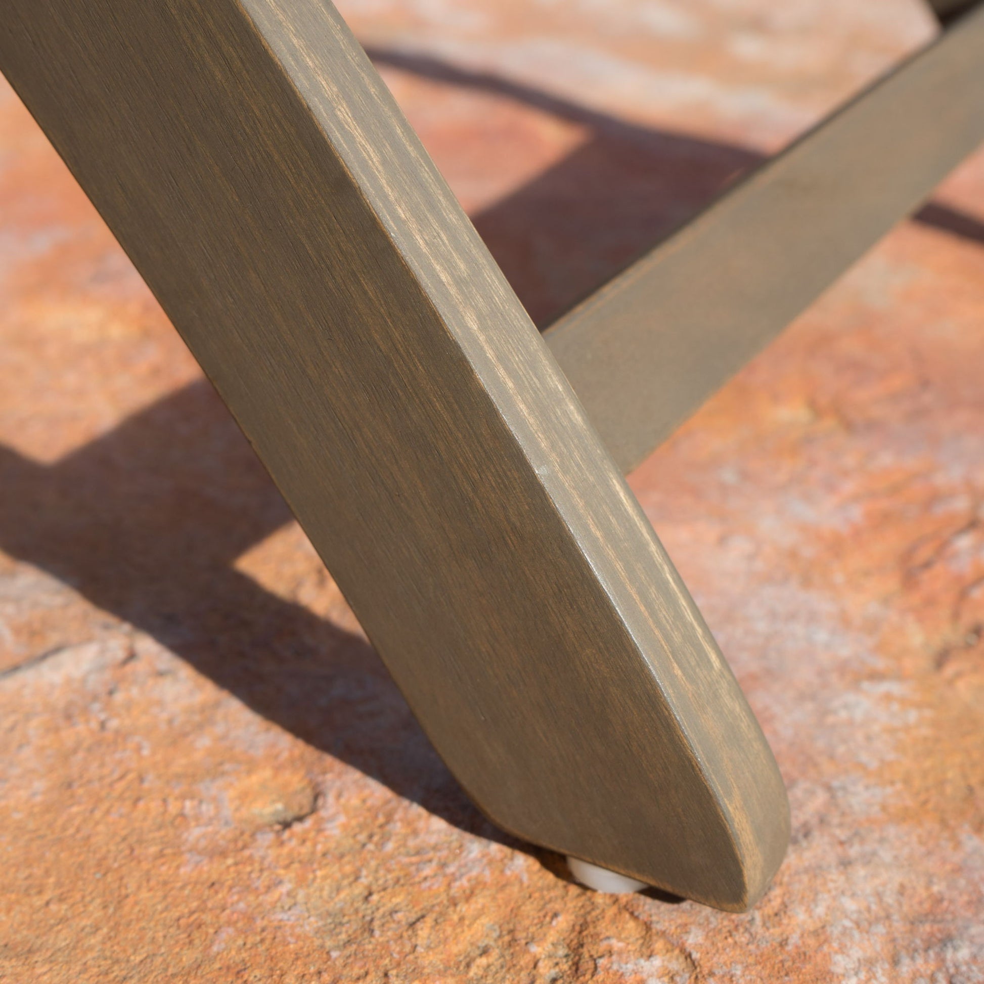 A close-up of the Positano - Foldable Dining Set shows its smooth, angled wooden leg resting on a textured, reddish-brown stone surface.