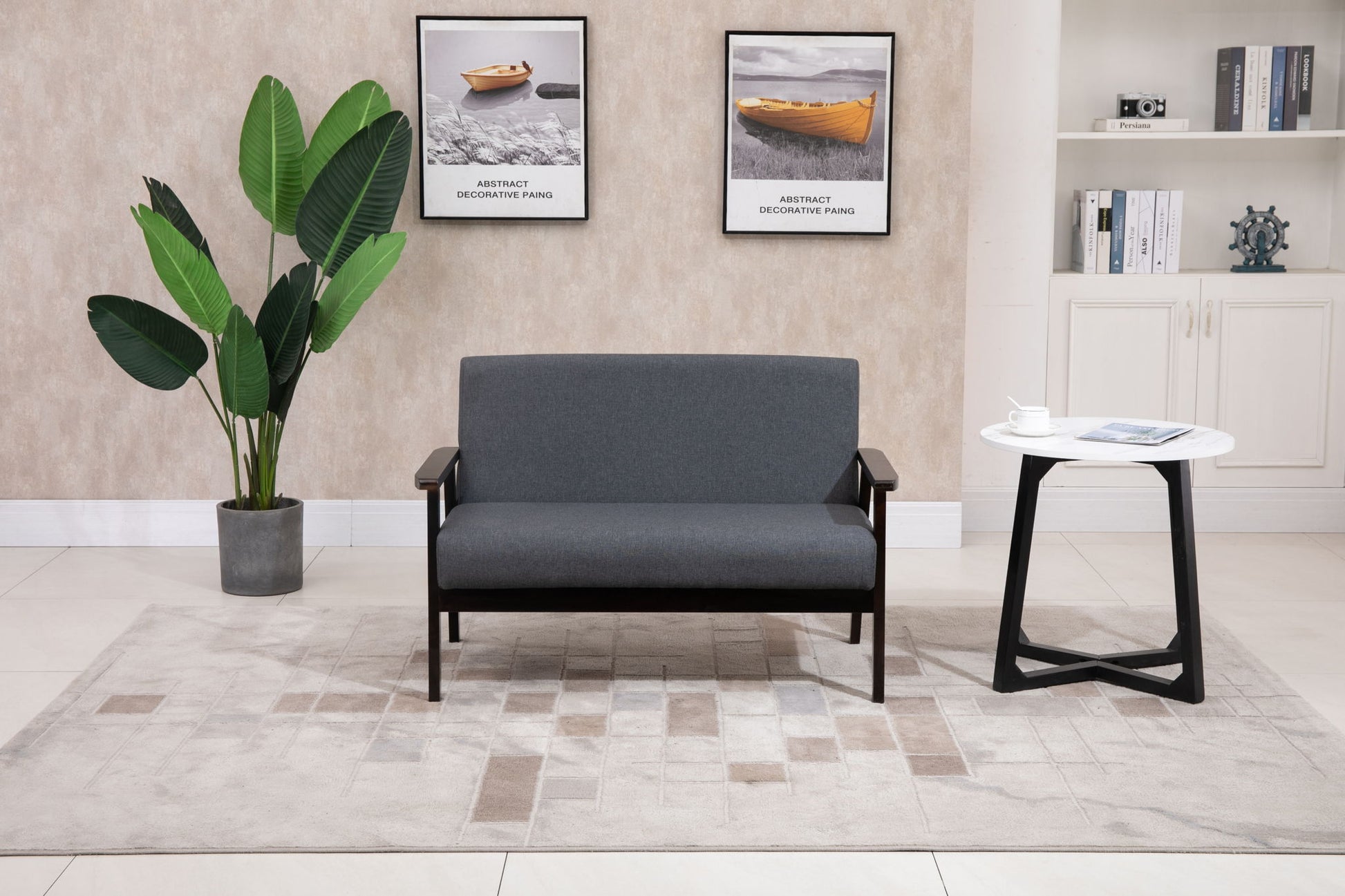 A modern living room featuring the Bahamas - Loveseat in gray, a potted plant, a white side table with a magazine and cup, boat-themed wall art, and a bookshelf in the background.