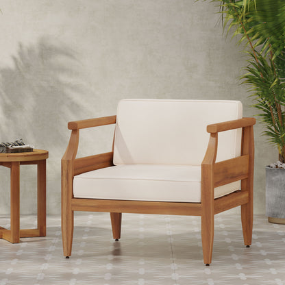 An Outdoor Club Chair With Recessed Arms And Cushions sits on a tiled floor beside a small wooden side table and a tall potted plant, placed against a light-colored wall.