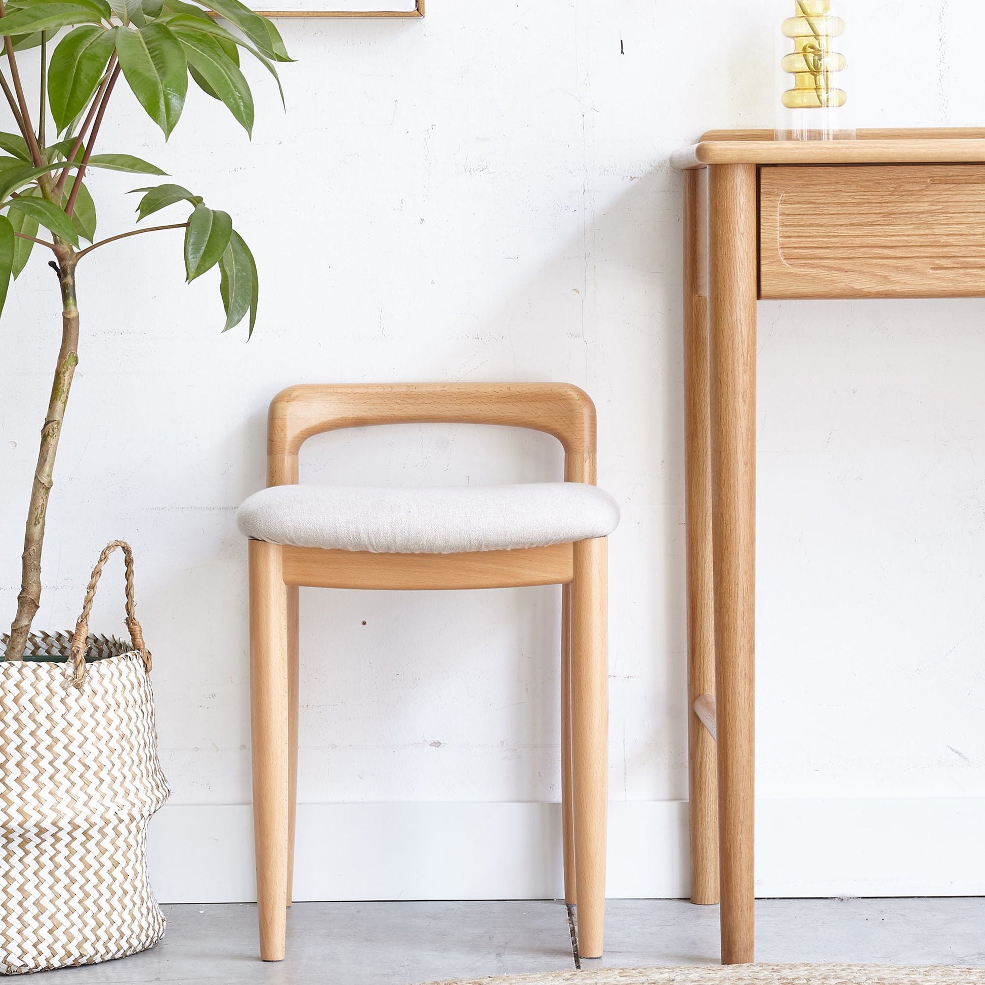 The Stool Bed For Dining Room - Natural, featuring light wood and a cushioned seat, sits by a wooden desk near a potted plant in a woven basket, set against white walls and a light floor.