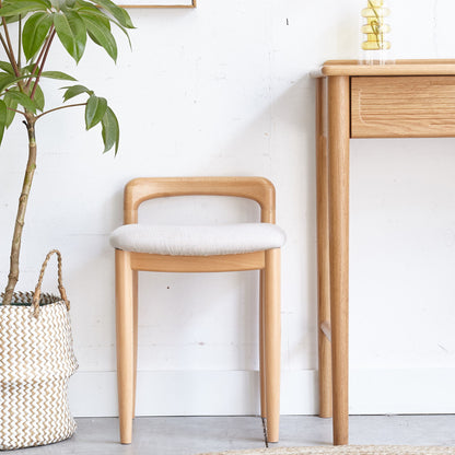 The Stool Bed For Dining Room - Natural, featuring light wood and a cushioned seat, sits by a wooden desk near a potted plant in a woven basket, set against white walls and a light floor.