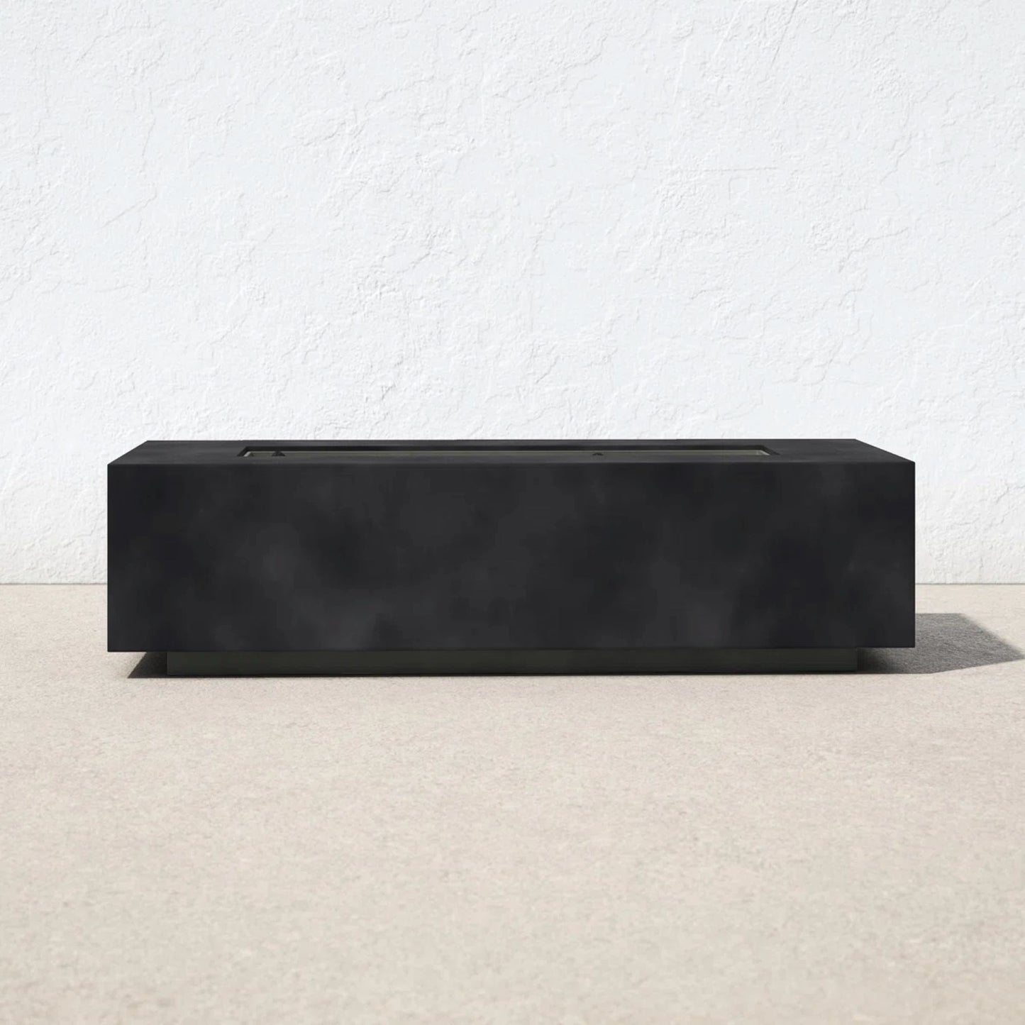A Modern Concrete Design Propane Outdoor Fire Pit Table with a sleek, rectangular black finish sits on a light outdoor surface against a white textured wall, featuring minimalist lines and a flat top.