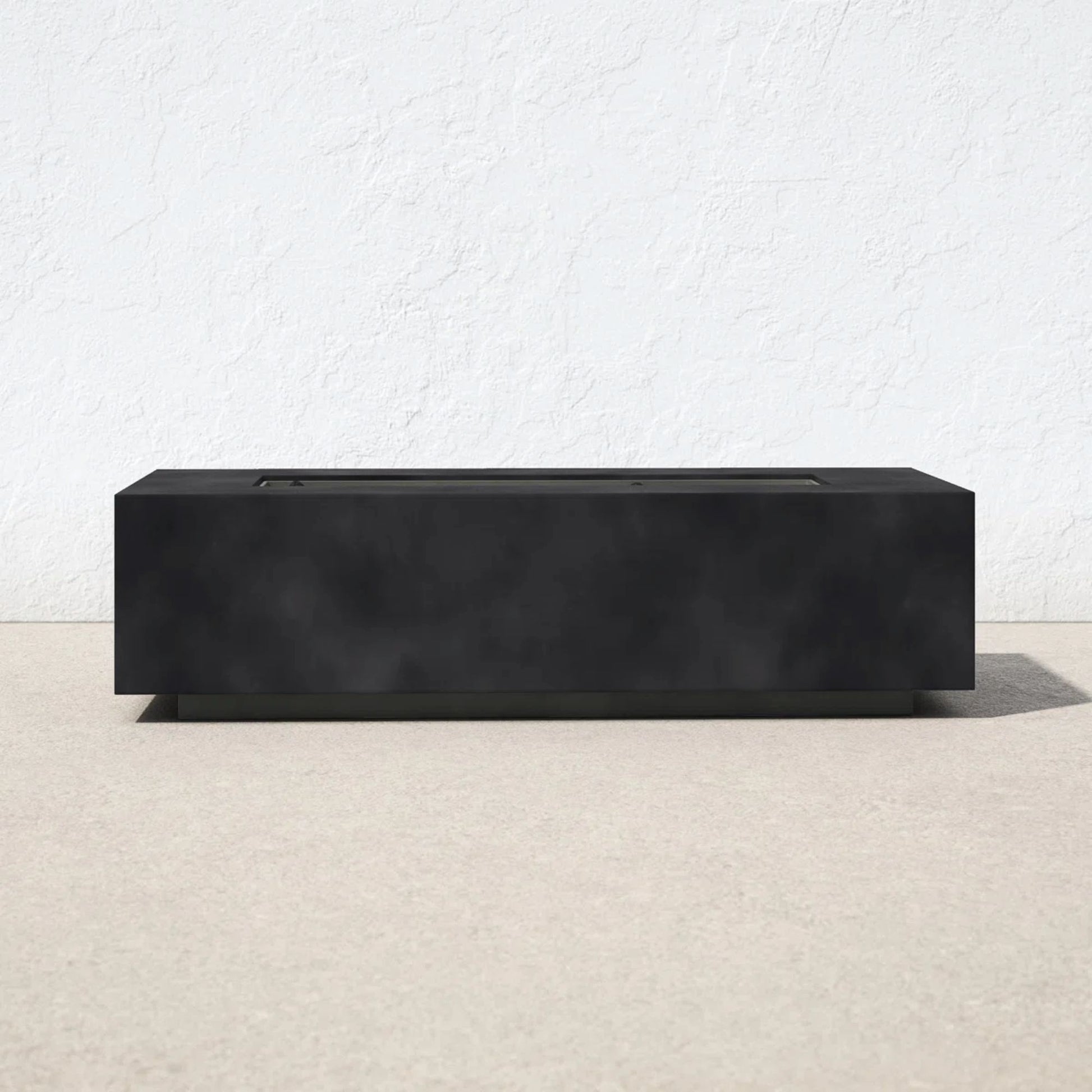 A Modern Concrete Design Propane Outdoor Fire Pit Table with a sleek, rectangular black finish sits on a light outdoor surface against a white textured wall, featuring minimalist lines and a flat top.