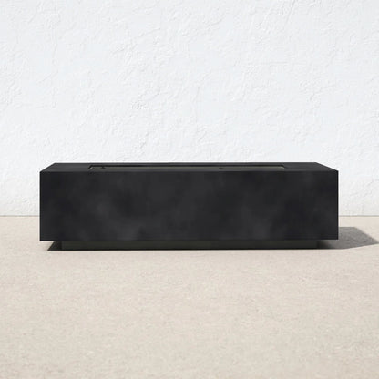 A Modern Concrete Design Propane Outdoor Fire Pit Table with a sleek, rectangular black finish sits on a light outdoor surface against a white textured wall, featuring minimalist lines and a flat top.
