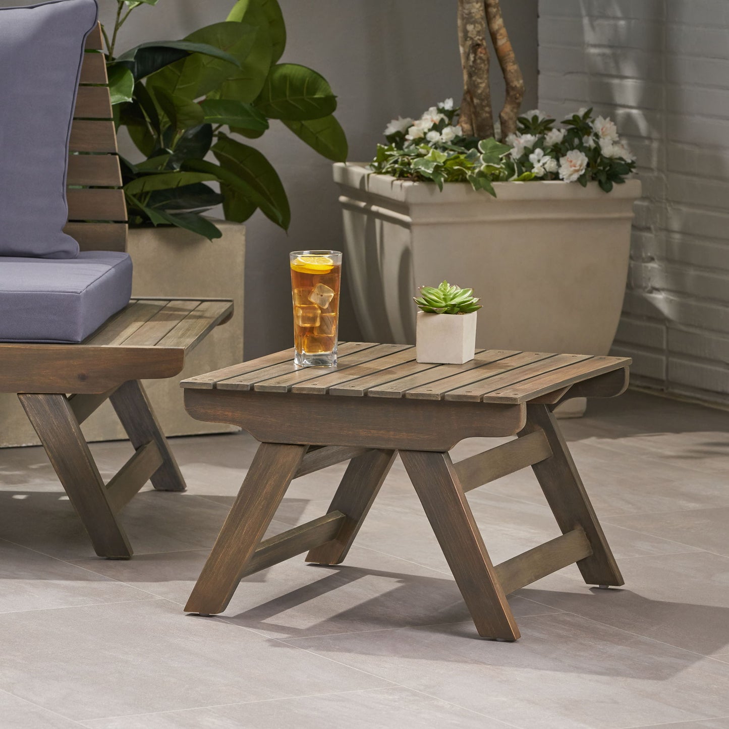 The Sedona Outdoor Side Table With Open Slat Design sits on a patio beside a chair, topped with a glass of iced tea and a small potted succulent. Green plants and white flowers in pots decorate the background.