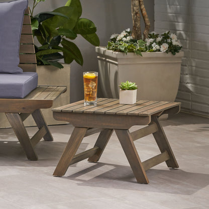 The Sedona Outdoor Side Table With Open Slat Design sits on a patio beside a chair, topped with a glass of iced tea and a small potted succulent. Green plants and white flowers in pots decorate the background.