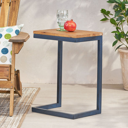 The C Shaped End Table With Iron Base And Fir Wood Top holds a glass of water and a pomegranate. It's next to a wooden chair featuring a colorful dotted pillow, with a potted plant placed close by.