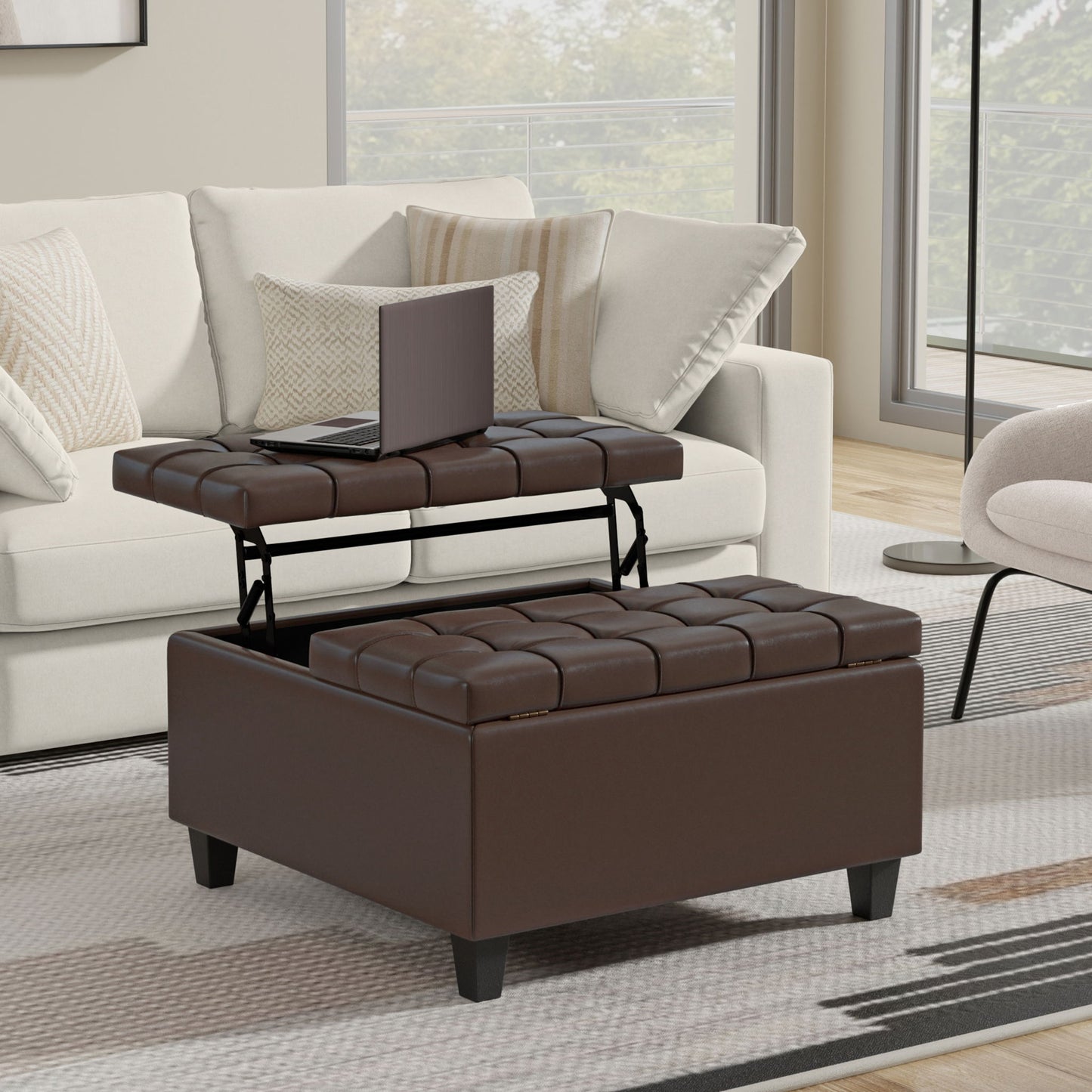 The Harrison - Large Coffee Table Storage Ottoman with a lift-top tray holding a laptop sits in front of a beige sofa with patterned pillows in a modern living room with large windows and a light rug.