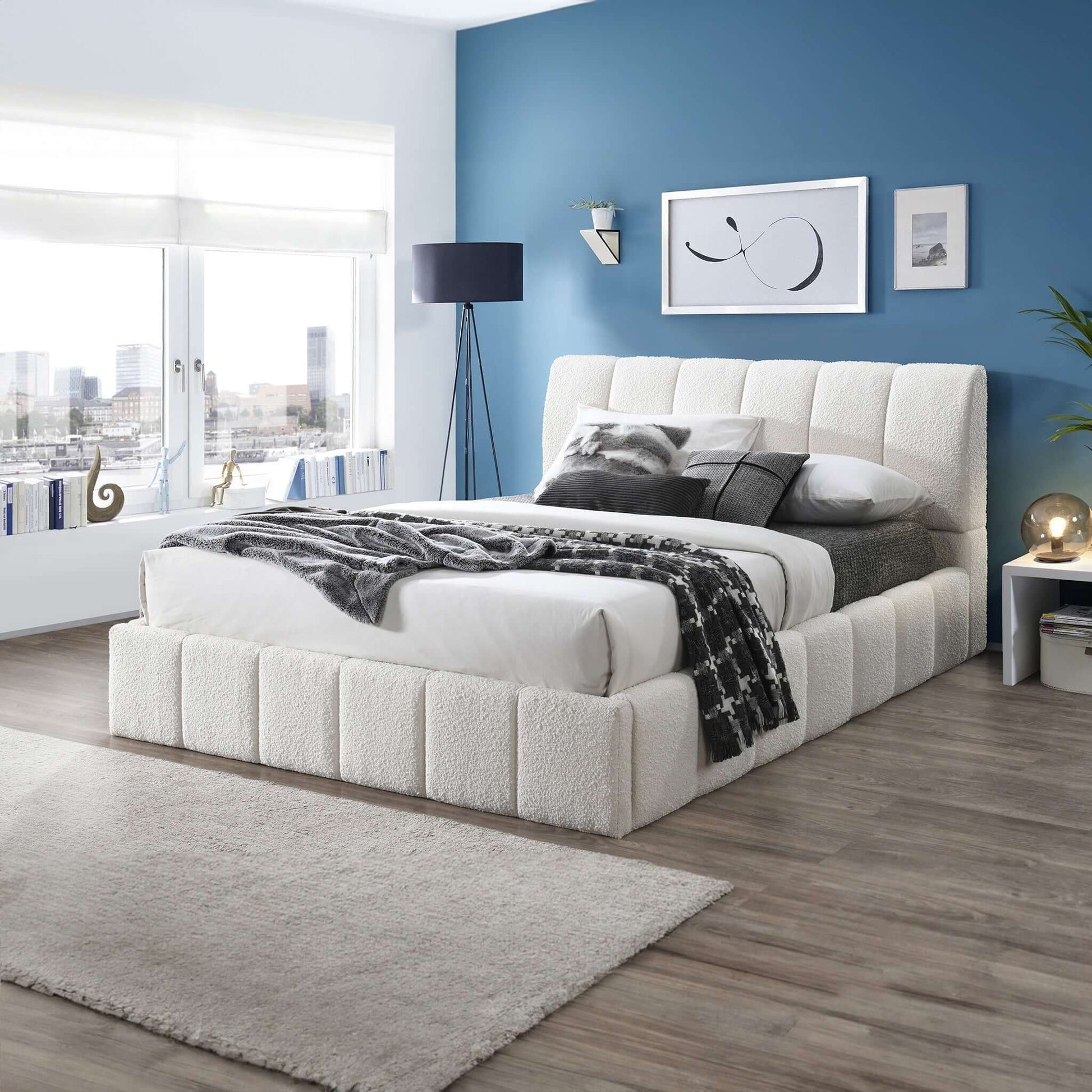 A modern bedroom features blue walls, a large window with city views, the Hilar Elegant Design Platform Bed in white with gray and white bedding, a nightstand, lamp, framed art, and a light gray rug over wood flooring.