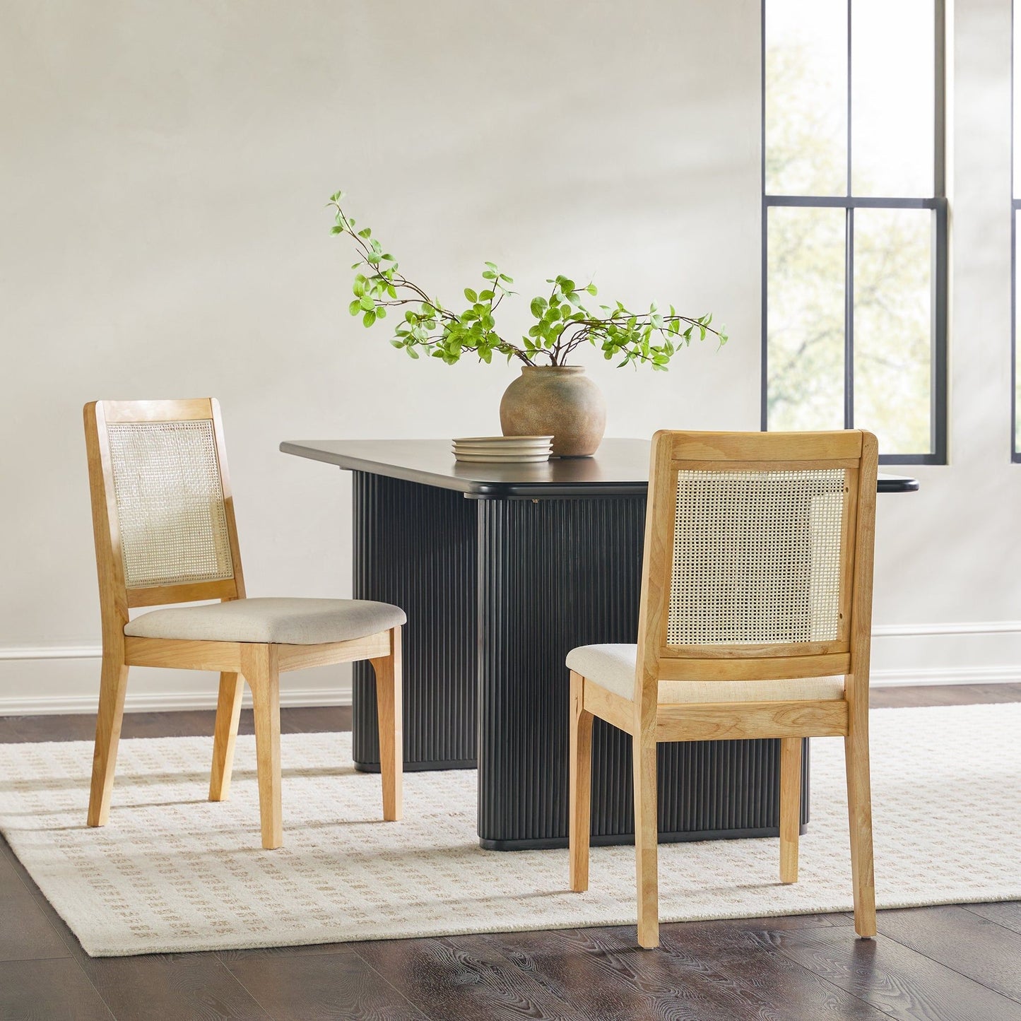 A minimalist dining area features a black rectangular table, a light rug, and a ceramic vase with green branches, paired with two Modern Solid Wood Dining Chairs With Rattan Inset Back (Set of 2), by large windows with natural light.