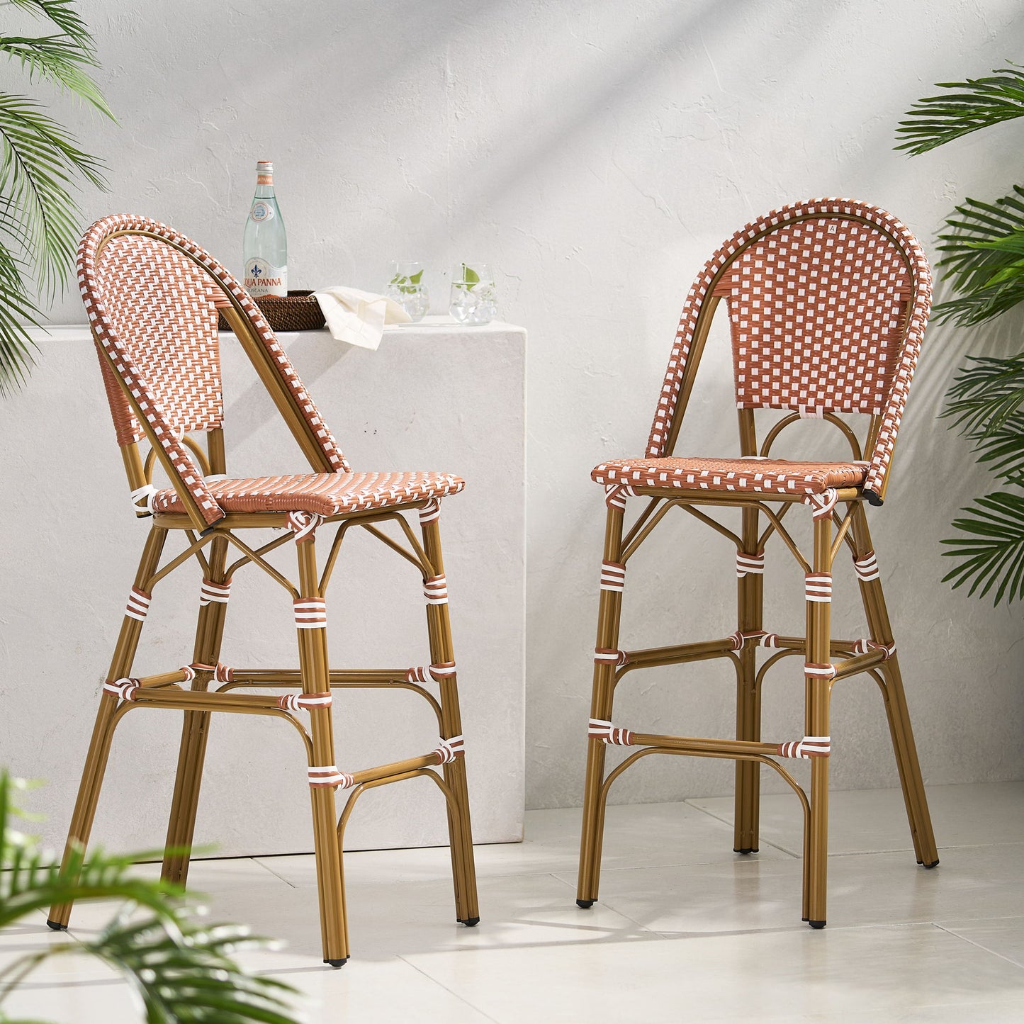 Two Outdoor Pe Rattan French Barstools with brown and white checkered seats and backs are placed by a white stone pedestal adorned with glasses and a bottle, while green plants frame the sunlit space.