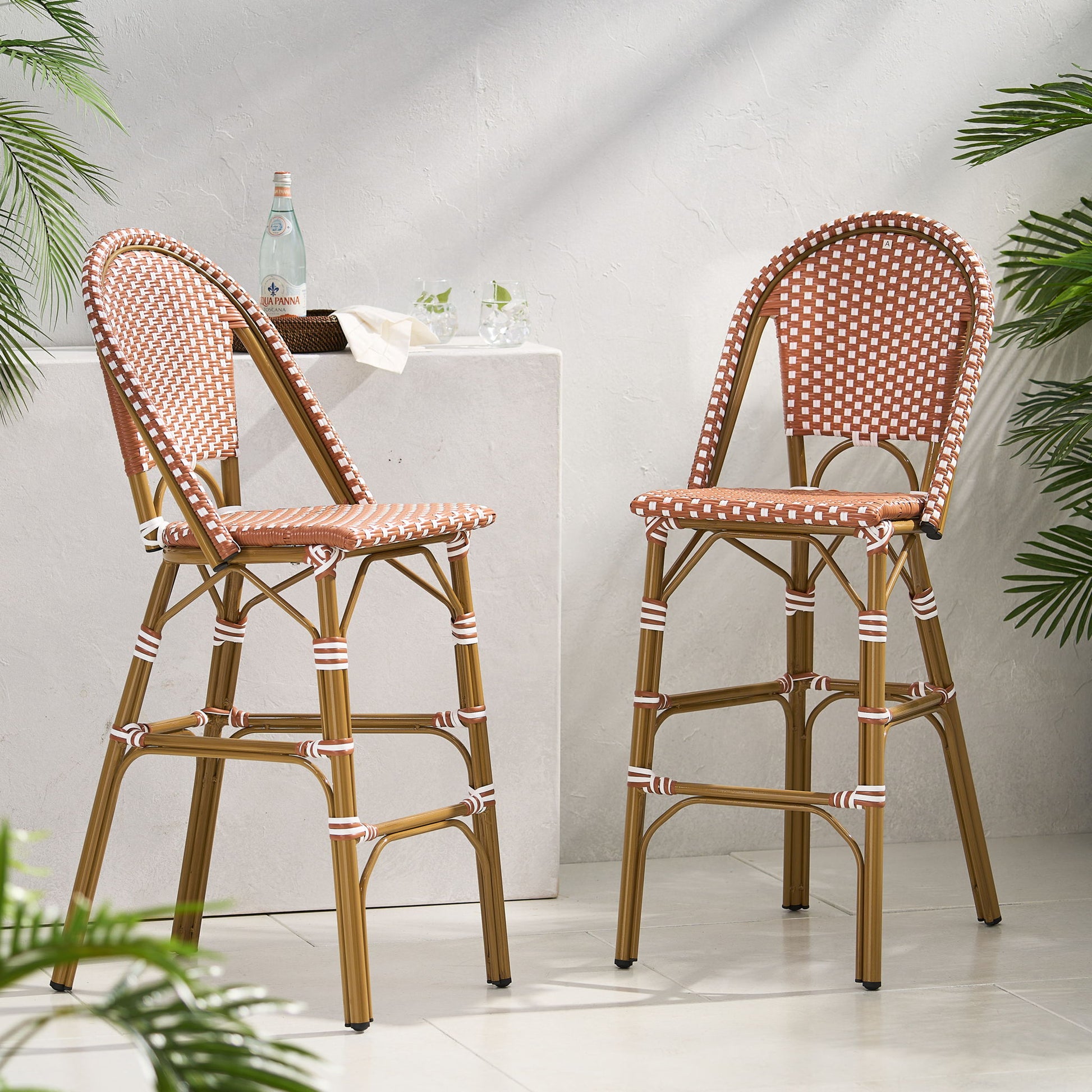 Two Outdoor Pe Rattan French Barstools with brown and white checkered seats and backs are placed by a white stone pedestal adorned with glasses and a bottle, while green plants frame the sunlit space.