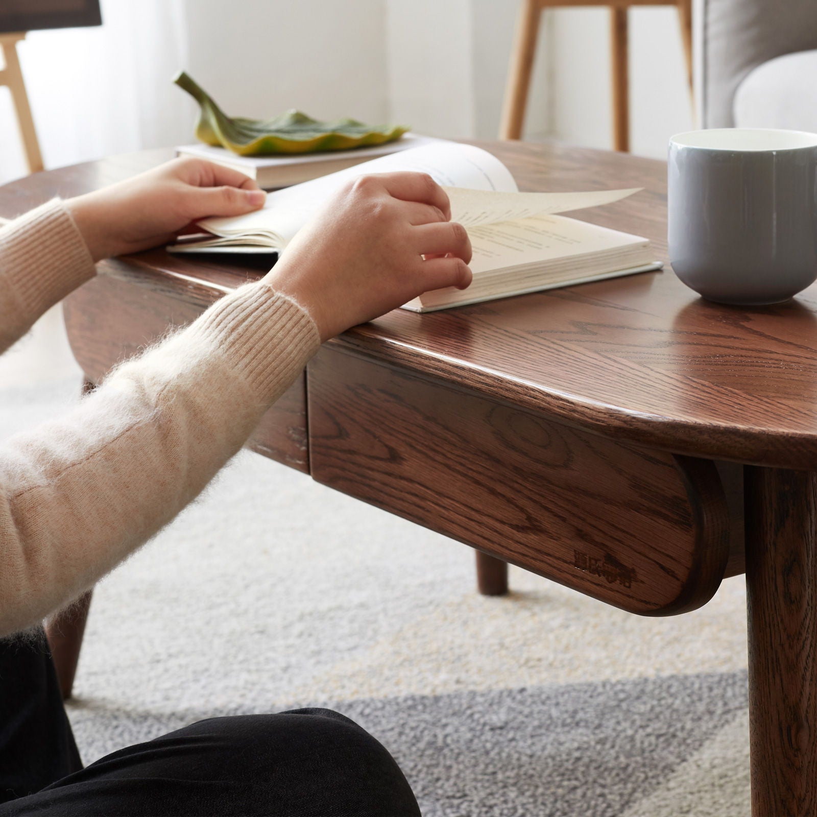 A person sits at the Capsule Centre Low Table With Drawers Coffee Table, reading an open book. A gray mug and a green decorative dish rest on the table. The cozy scene features soft lighting and carpeted flooring.