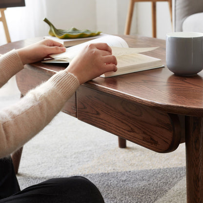A person sits at the Capsule Centre Low Table With Drawers Coffee Table, reading an open book. A gray mug and a green decorative dish rest on the table. The cozy scene features soft lighting and carpeted flooring.